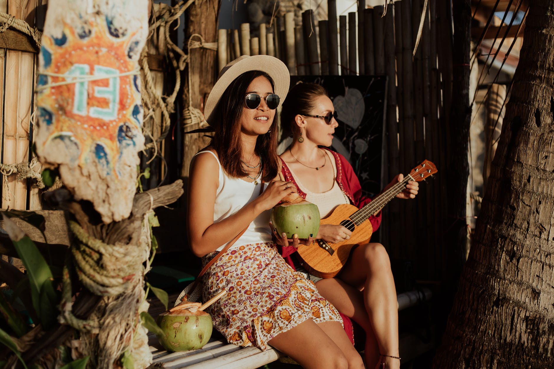 women sitting on a bamboo bench while playing guitar and drinking coconut juice