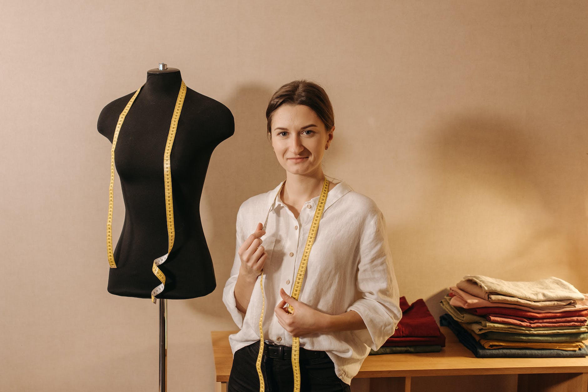 dressmaker smiling in white long sleeve blouse standing beside black manikin with measuring tape hanging on her neck
